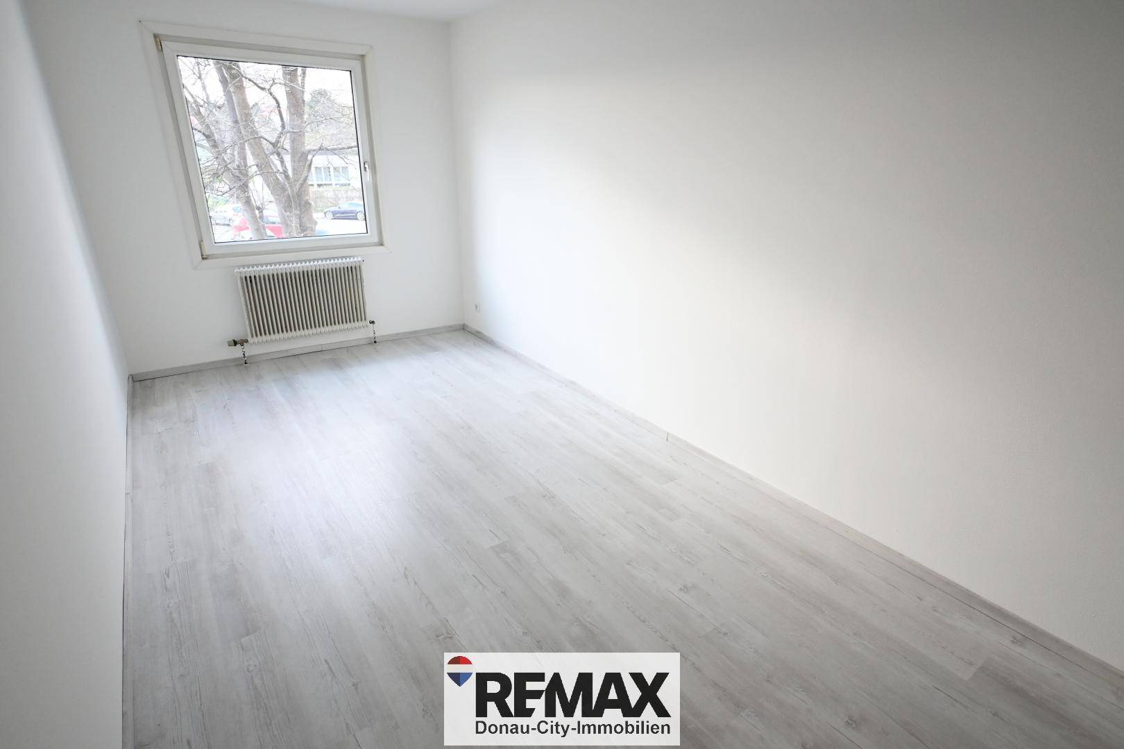 Helles Zimmer mit großem Fenster, hellem Laminatboden und weiß gestrichenen Wänden.