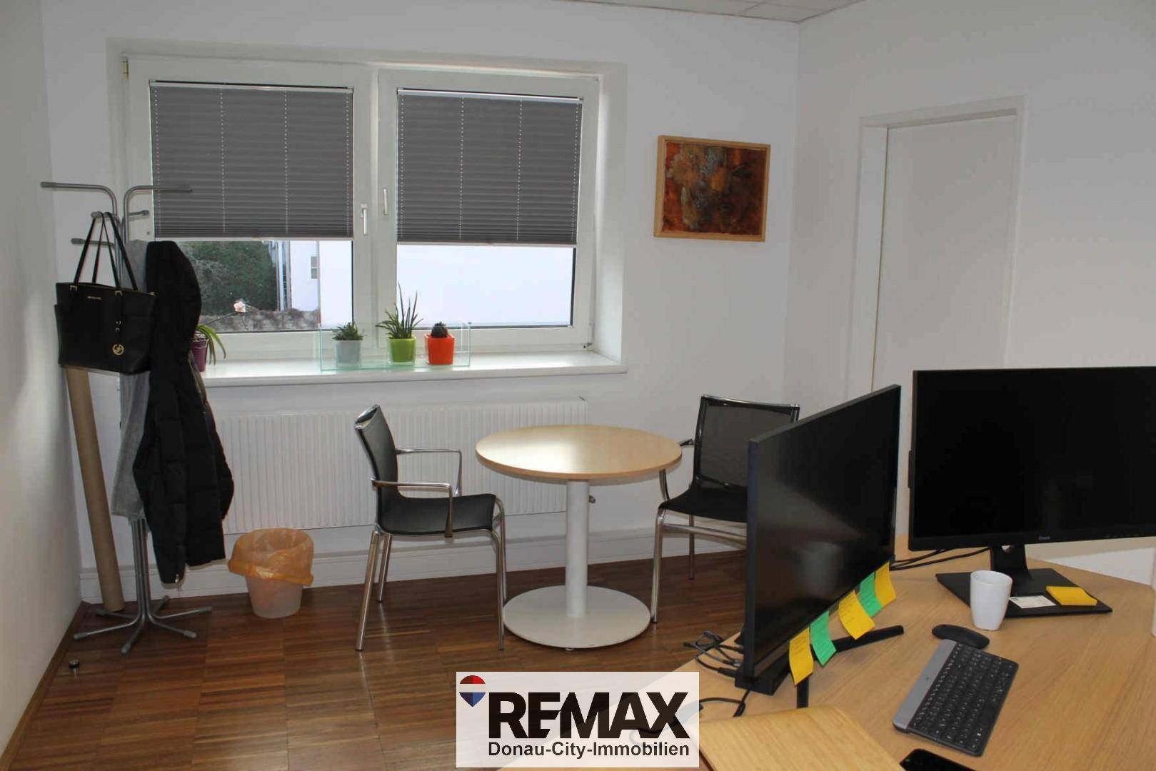 Kleiner Bürobereich mit Schreibtisch, Computermonitoren und einem runden Besprechungstisch am Fenster.