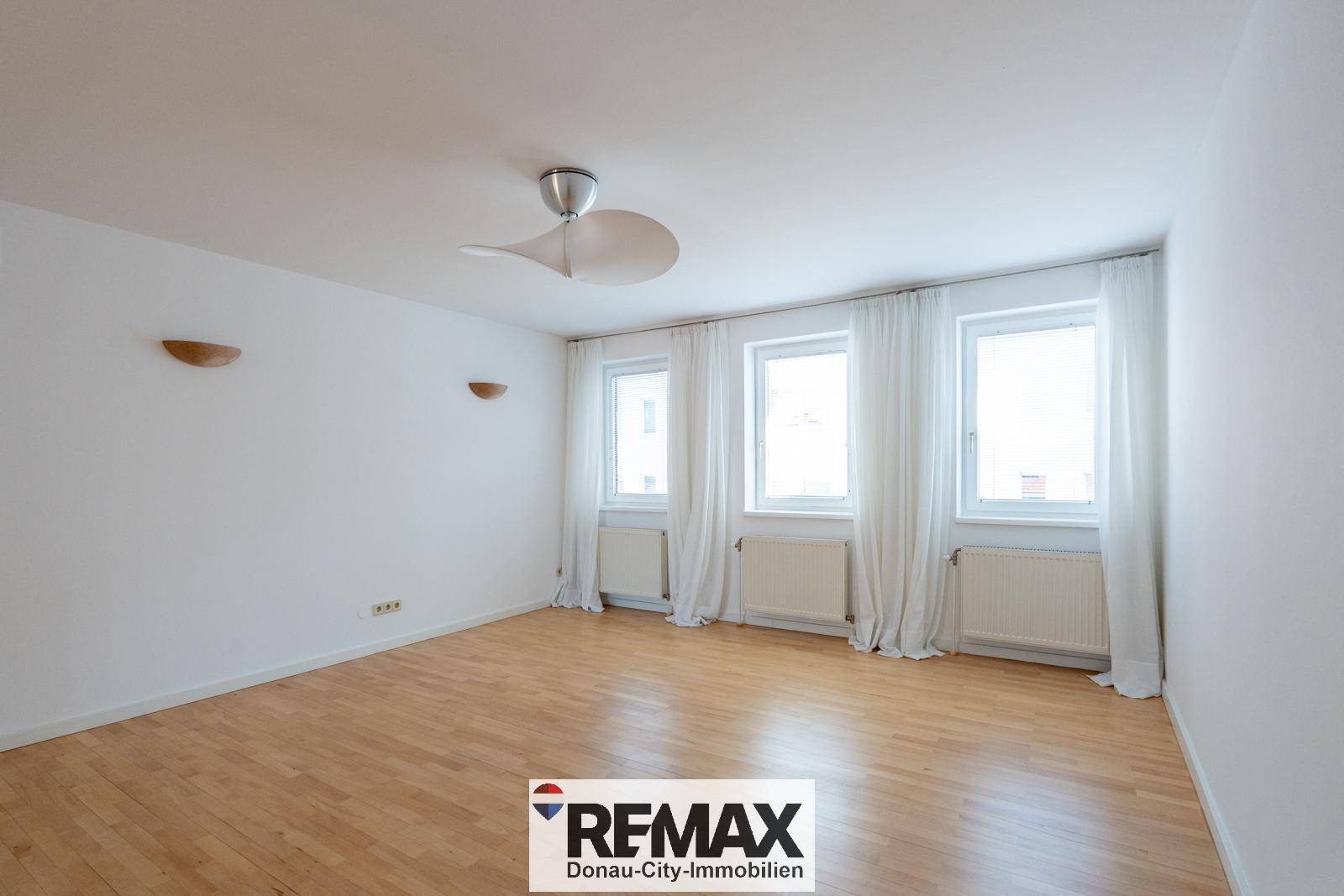 Helles Schlafzimmer mit Parkettboden, drei Fenstern und Deckenventilator.