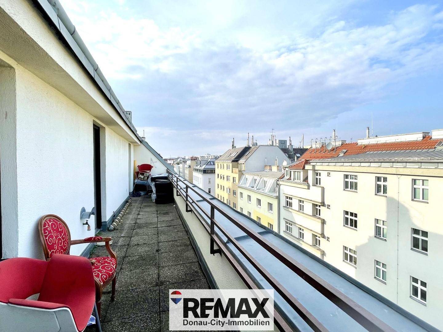 Langer Balkon mit Metallgeländer und Sitzgelegenheiten, bietet einen Blick auf die umliegenden Gebäude.