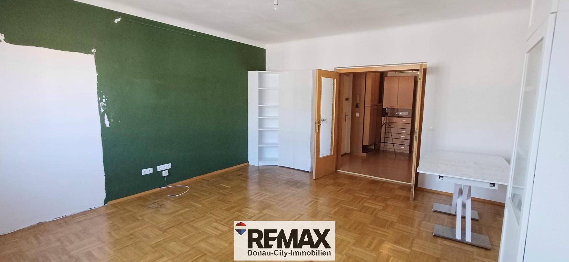 Heller Wohnraum mit Parkettboden, grüner Akzentwand und Zugang zu einem weiteren Zimmer.