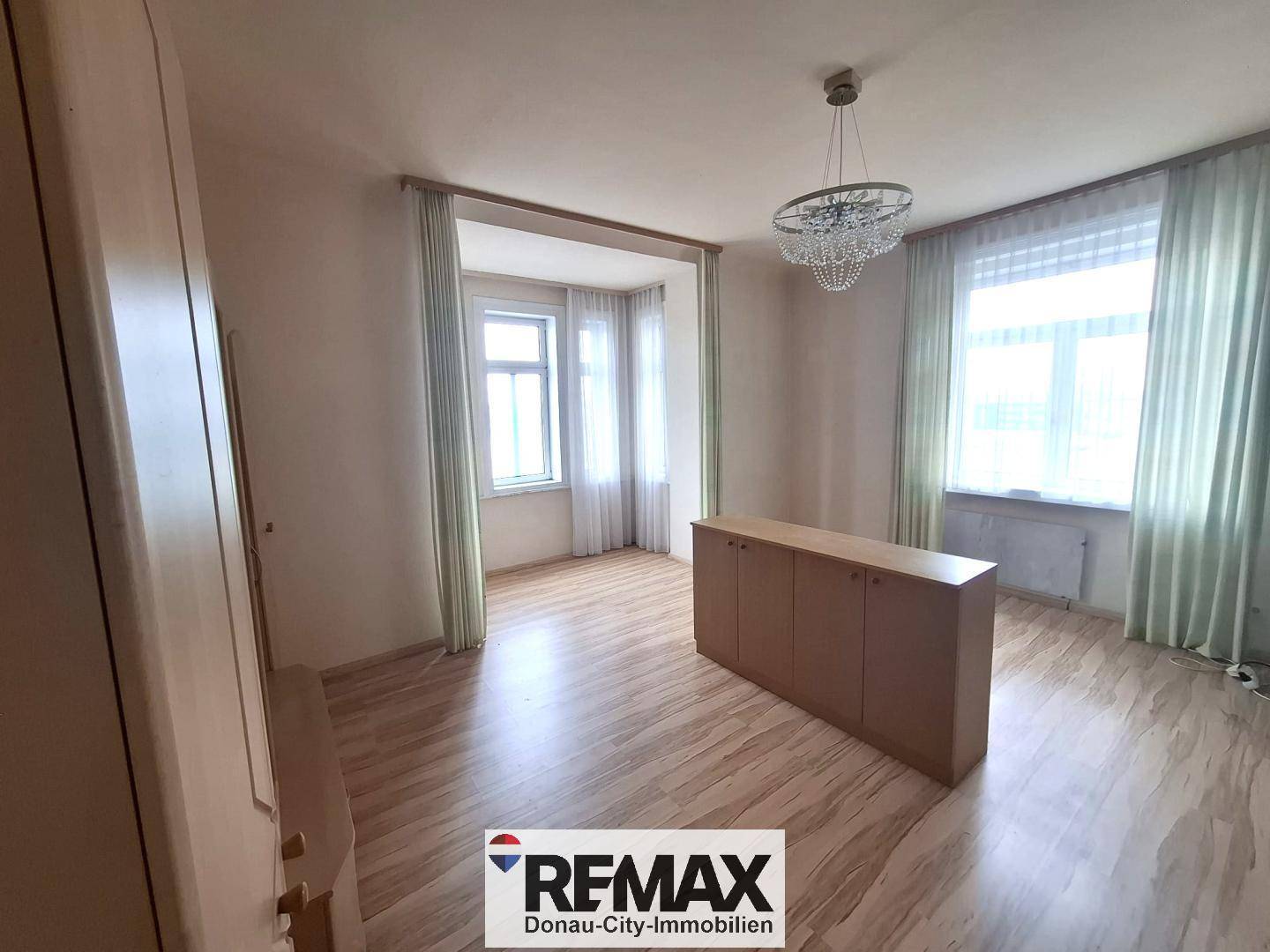 Heller Wohnraum mit Laminatboden, großen Fenstern und einem Kronleuchter.