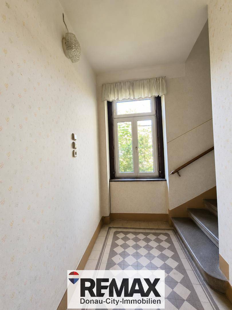 Treppenabsatz mit Fenster, gemusterter Tapete und Fliesenboden, der viel Tageslicht hereinlässt.