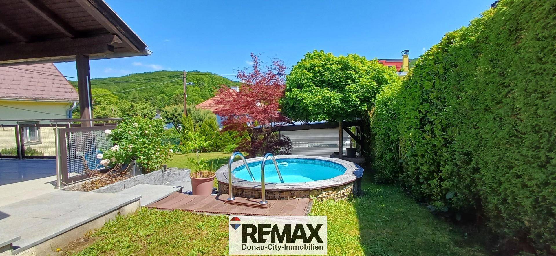 Kleiner runder Pool im Garten, umgeben von Holzdeck, Rasen und hohen Hecken.