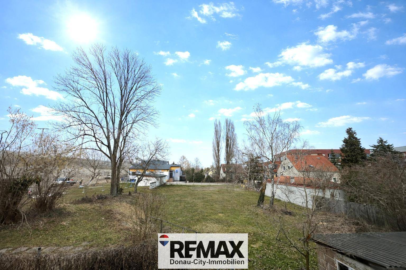 Weitläufiger Blick über eine grüne Wiese mit Bäumen und Häusern im Hintergrund.
