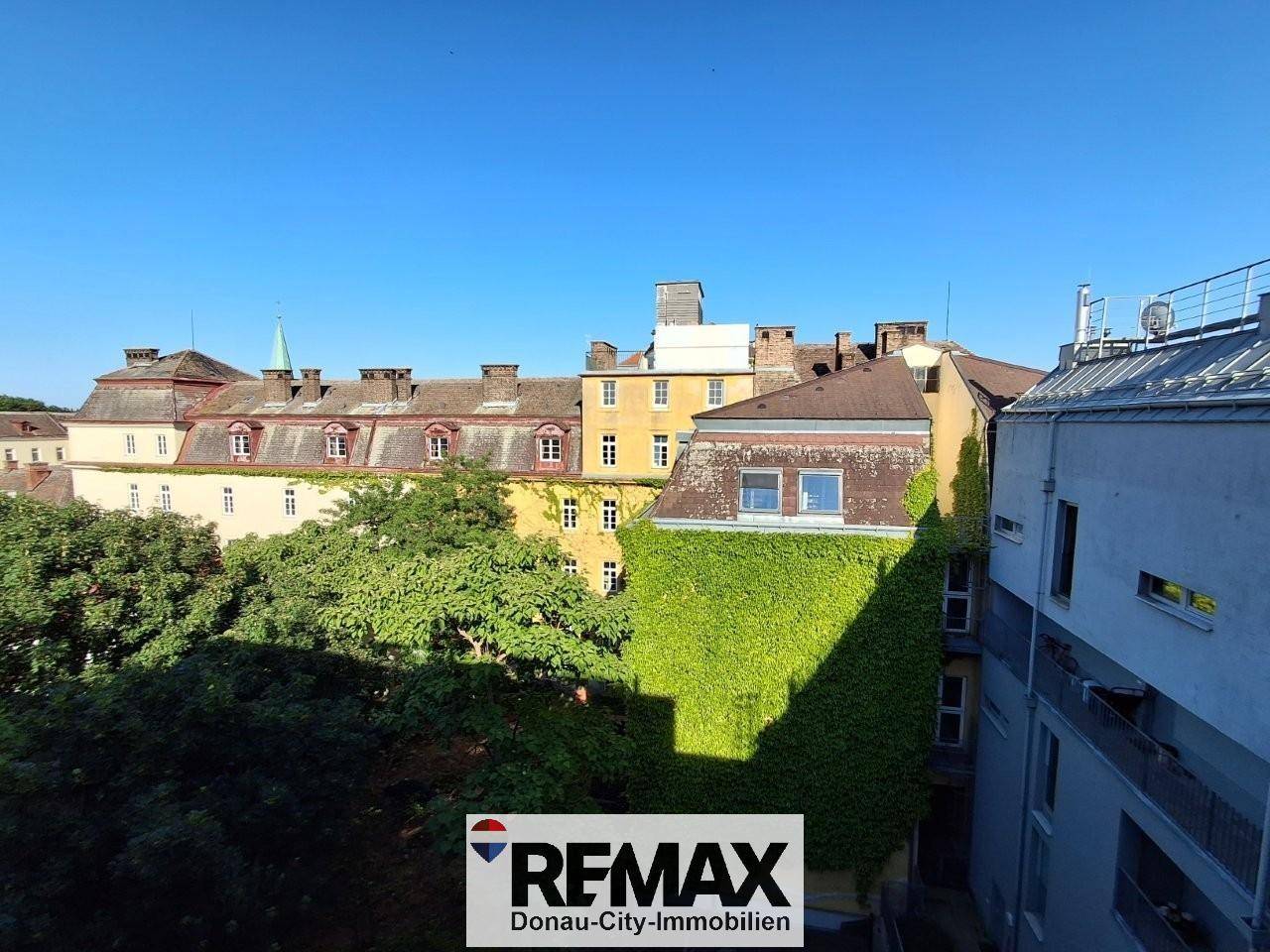 Historische Gebäude mit Efeubewuchs und grünen Bäumen im Vordergrund, unter blauem Himmel.