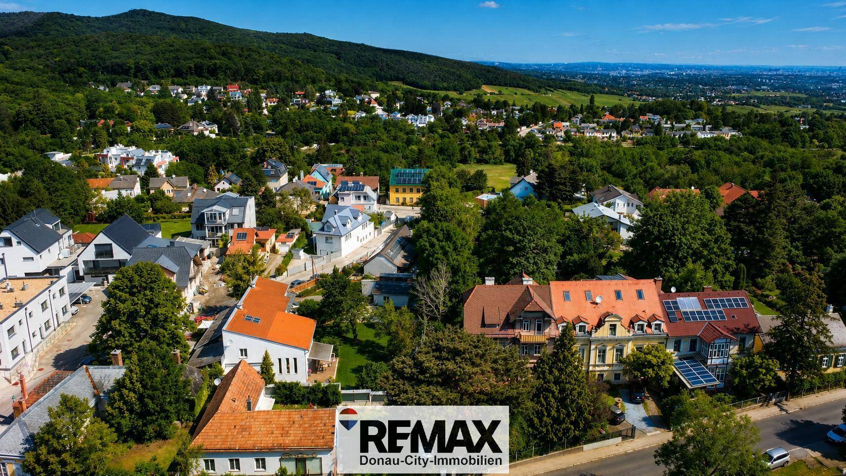 Panoramablick auf eine grüne Wohngegend mit verschiedenen Häusern und bewaldeten Hügeln im Hintergrund.