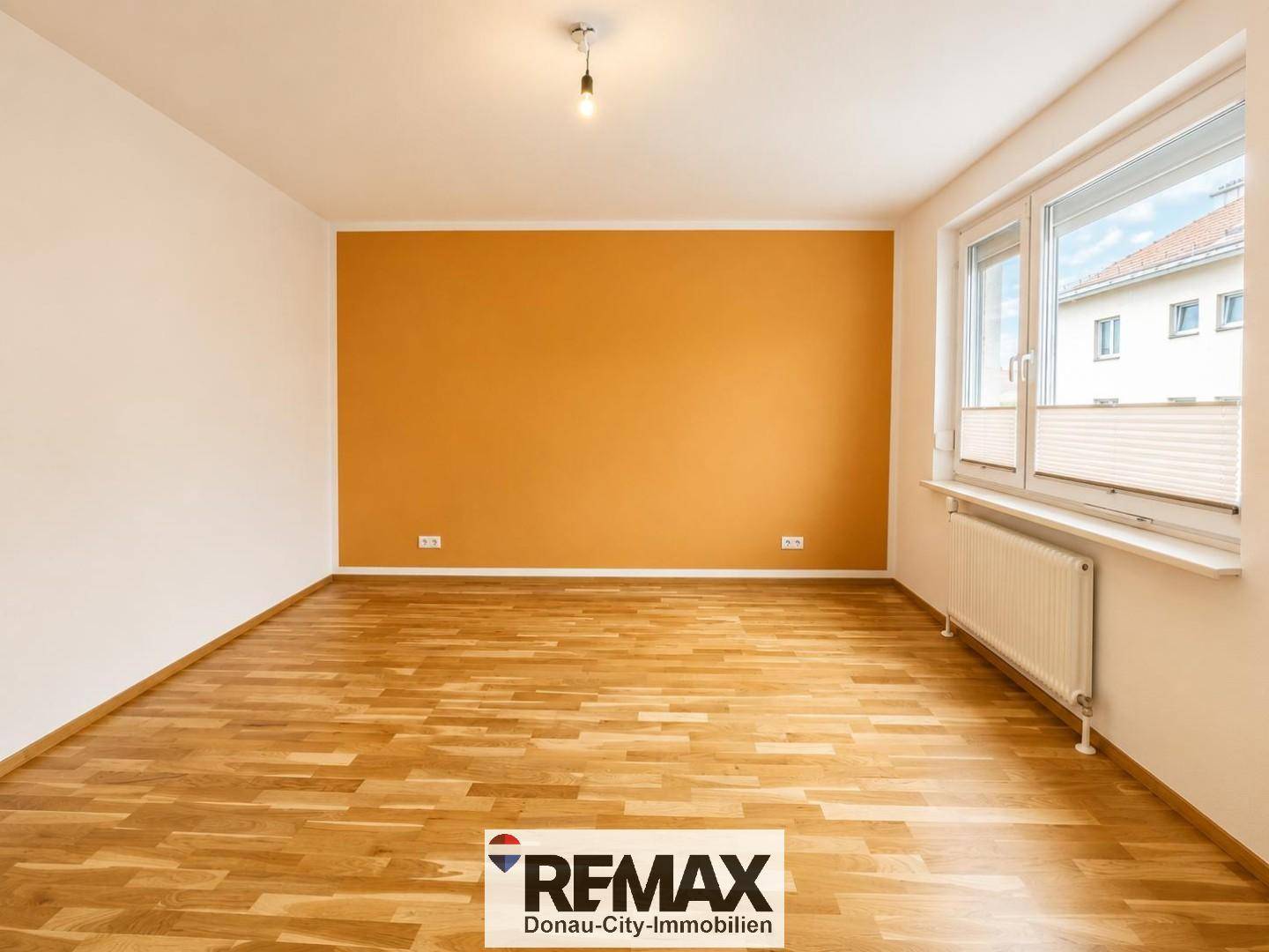 Geräumiges Wohnzimmer mit hellem Parkettboden und einer auffälligen orangefarbenen Akzentwand.