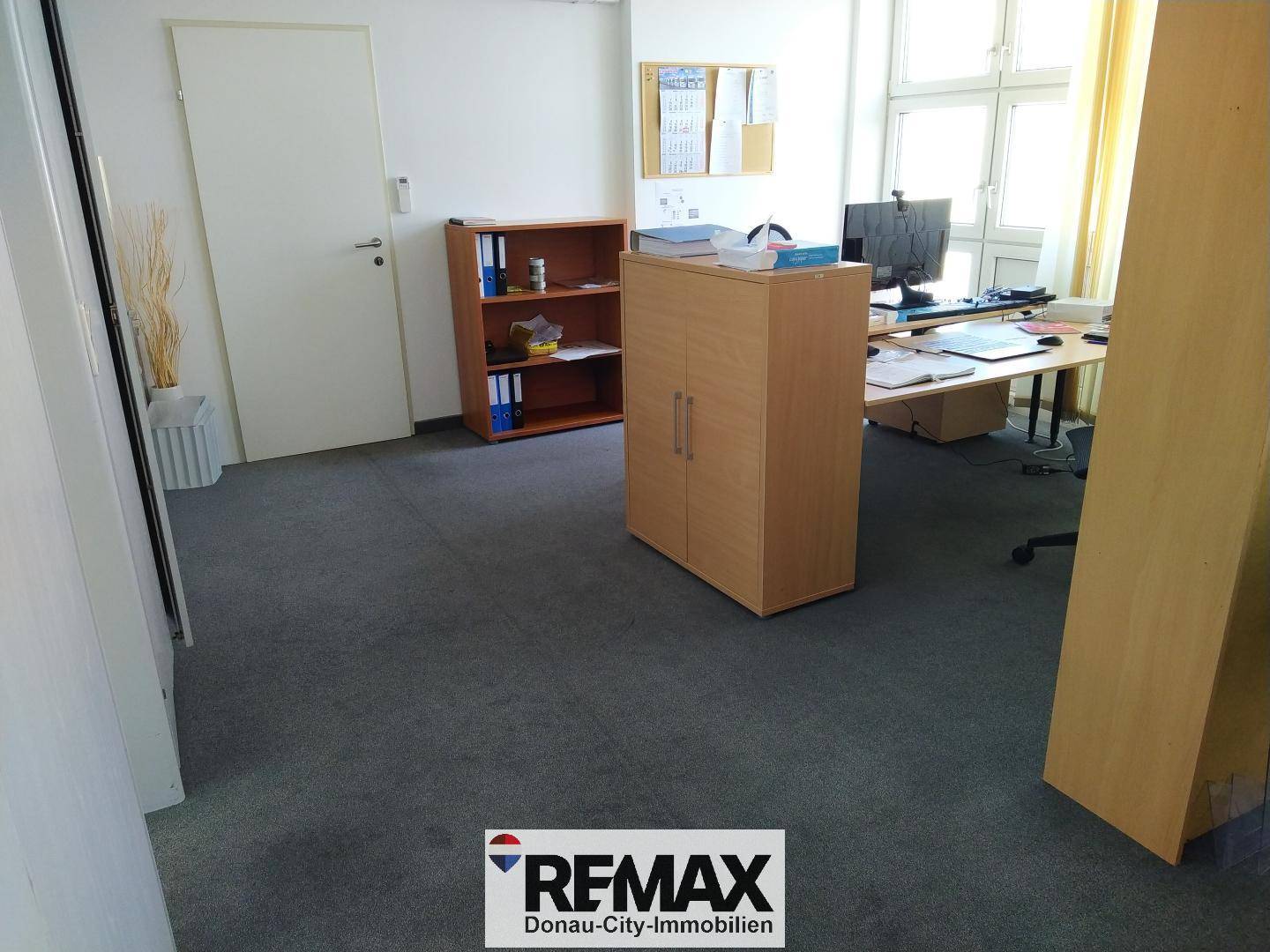 Geräumiges Büro mit Teppichboden, Holzregalen und einem Arbeitsplatz am Fenster für Tageslicht.