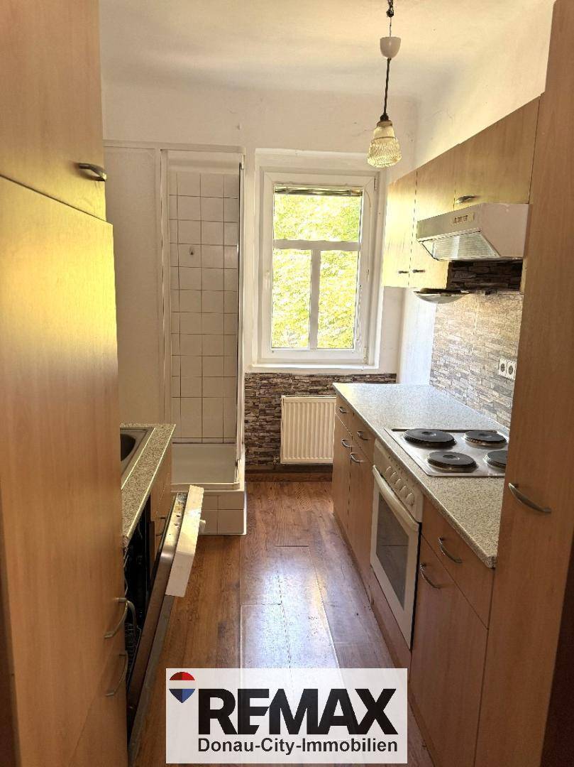 Schmale Küche mit Holzschränken, Fenster und integrierter Duschkabine am Ende des Raumes.