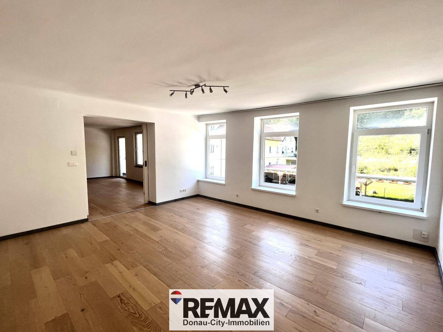 Geräumiger Wohnbereich mit Holzboden, mehreren Fenstern und Zugang zu einem weiteren Zimmer.