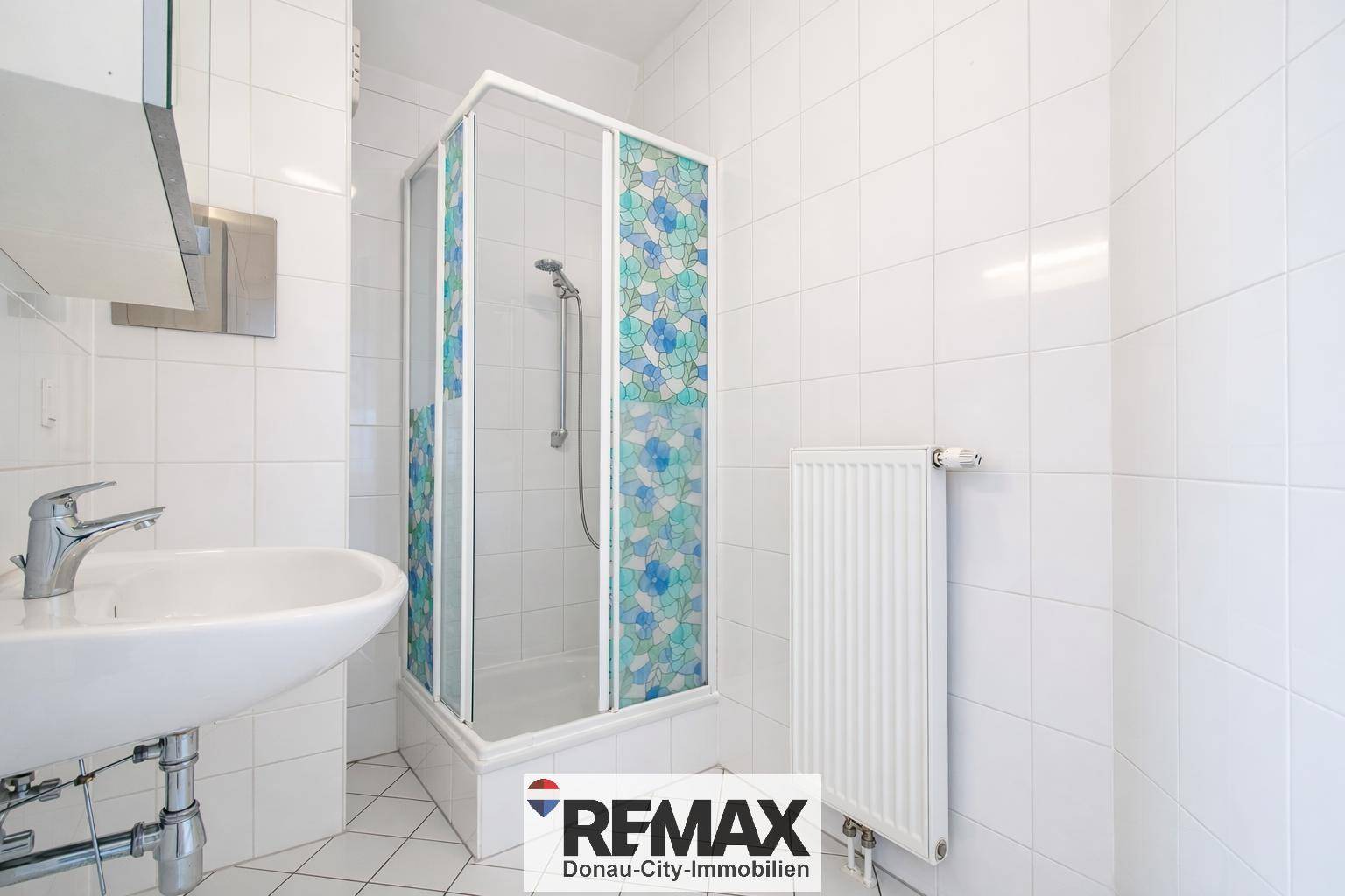 Funktionales Badezimmer mit weißen Fliesen, Einzelwaschbecken und Duschkabine mit gemustertem Glas.