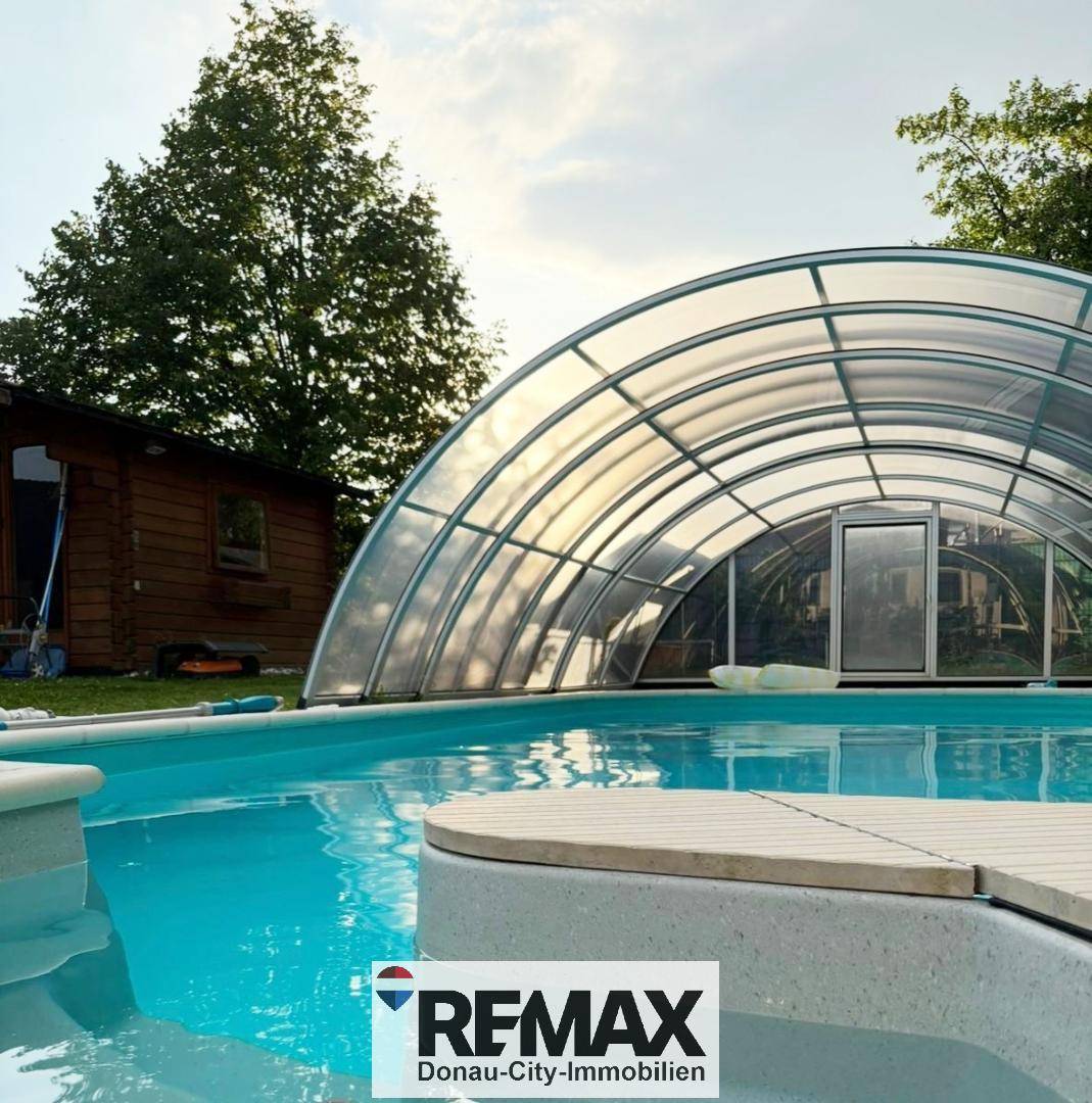 Überdachter Swimmingpool mit Holzdeck und angrenzendem Gartenhaus, umgeben von Bäumen.