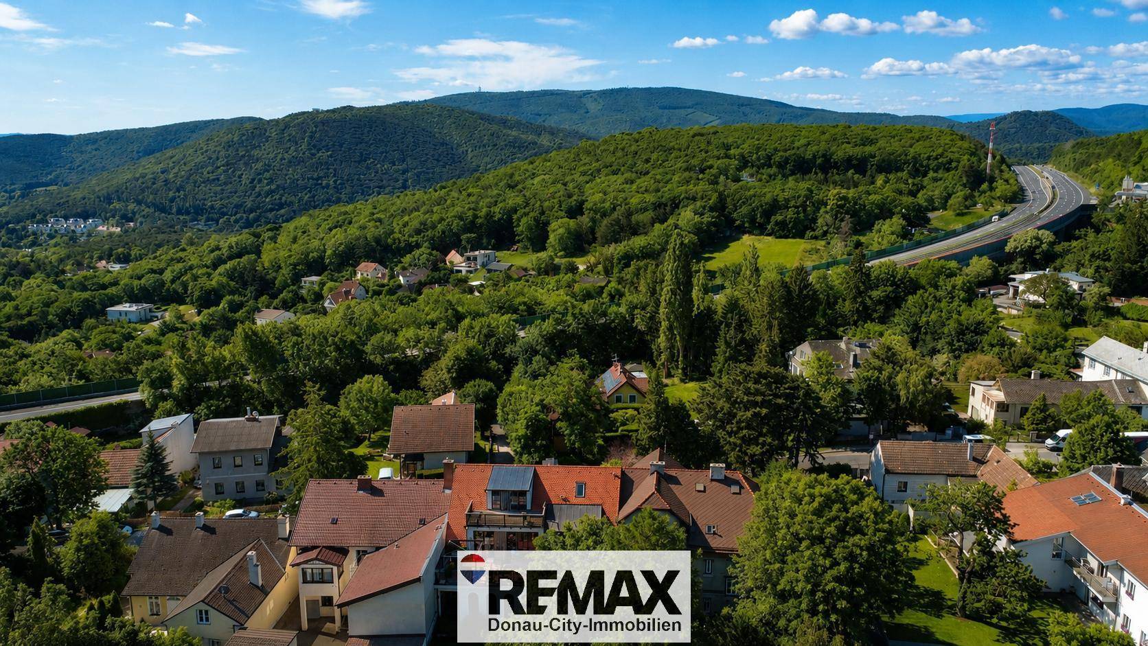 Panoramablick auf eine malerische Stadt in einem Tal, umgeben von bewaldeten Hügeln und einer Landstraße.