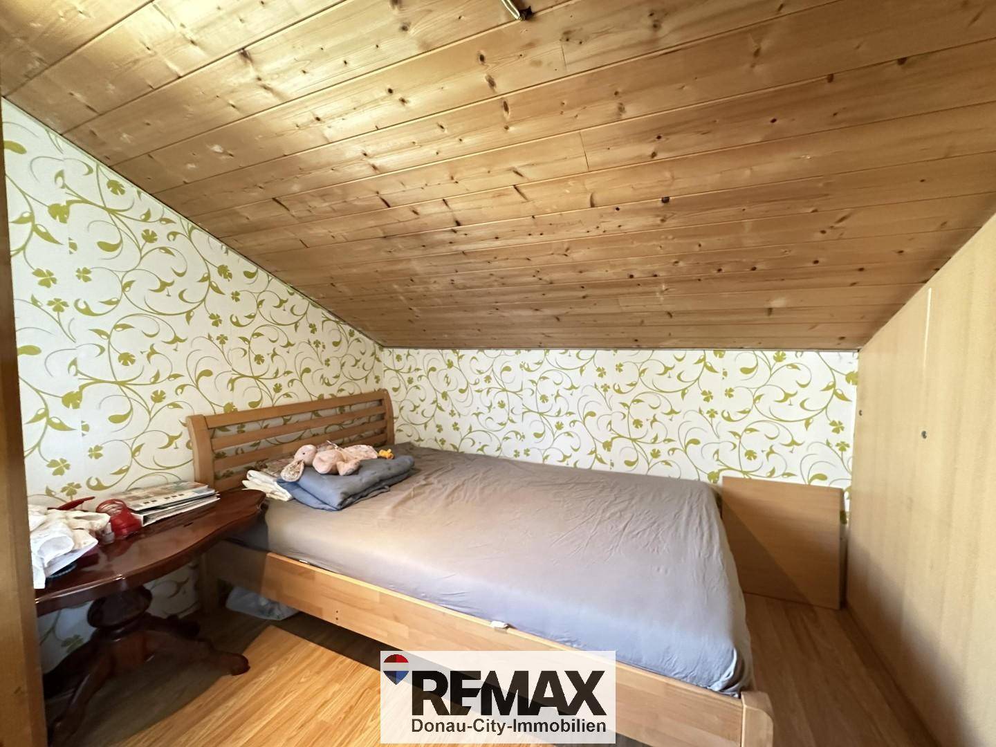 Schlafzimmer mit Holzdachschräge, gemusterter Tapete und einfachem Holzbett.