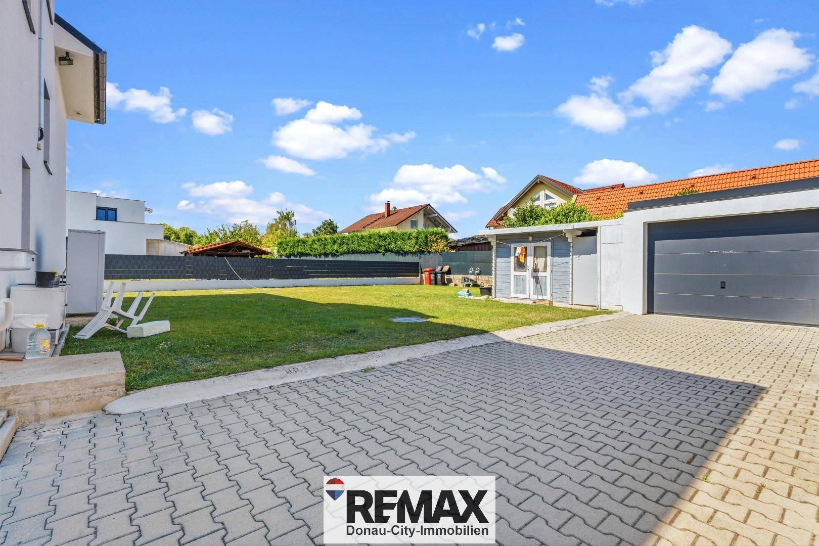 Großer, gepflegter Gartenbereich mit Rasenfläche, Gartenhaus und gepflasterter Zufahrt zur Garage.