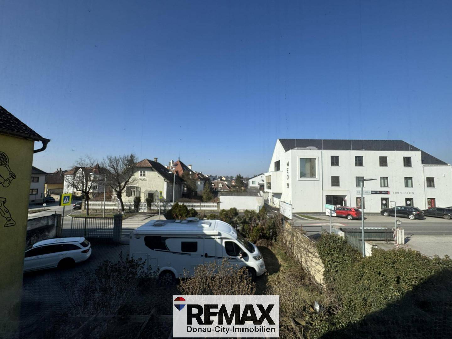 Blick aus dem Fenster auf die Umgebung mit einer Straße, geparkten Fahrzeugen und weiteren Gebäuden.