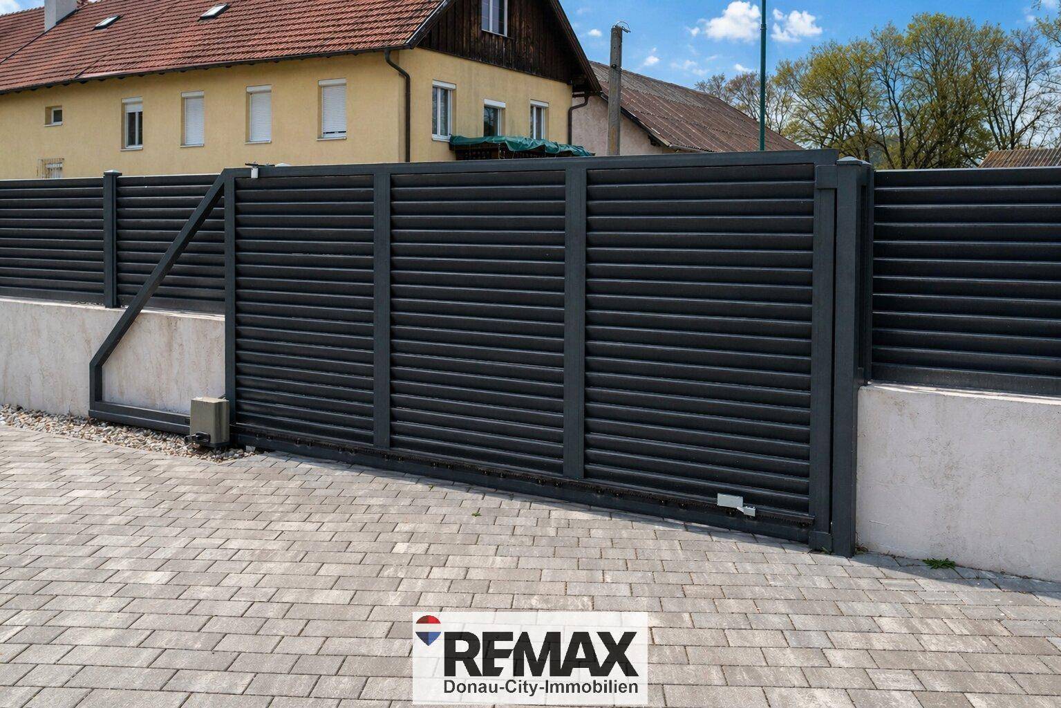 Moderne schwarze Zaunanlage mit Schiebetor und Pflastersteinen vor einem Wohnhaus.