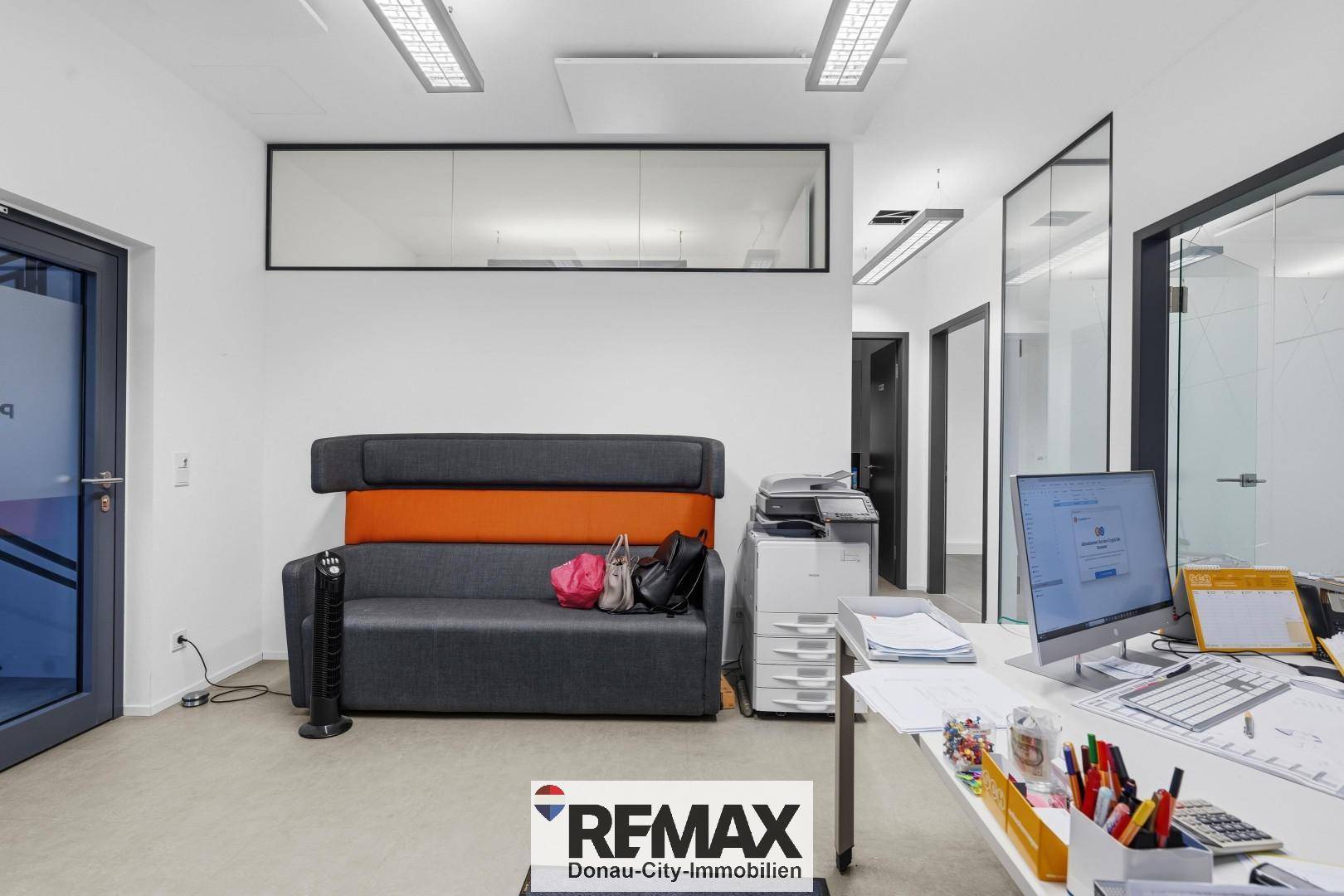 Büro mit Schreibtisch, Sofa und Drucker, durch Glastrennwand vom Flur getrennt.