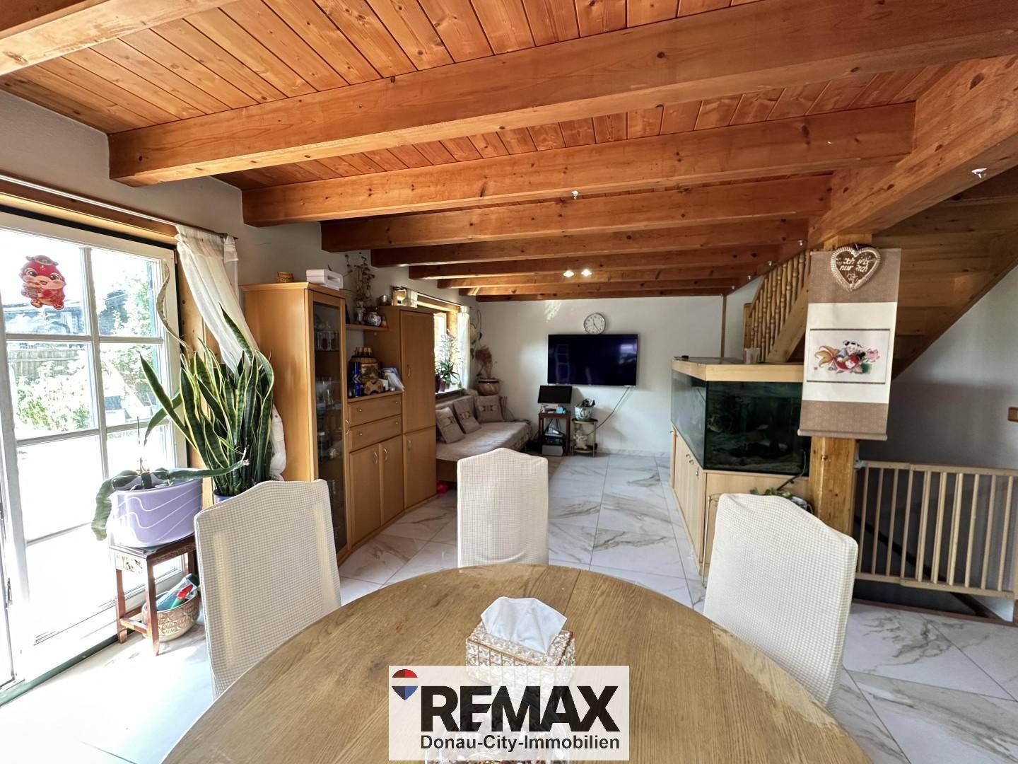 Heller Wohnbereich mit Esstisch, Sofa, Fernseher und großem Aquarium unter Holzbalkendecke.