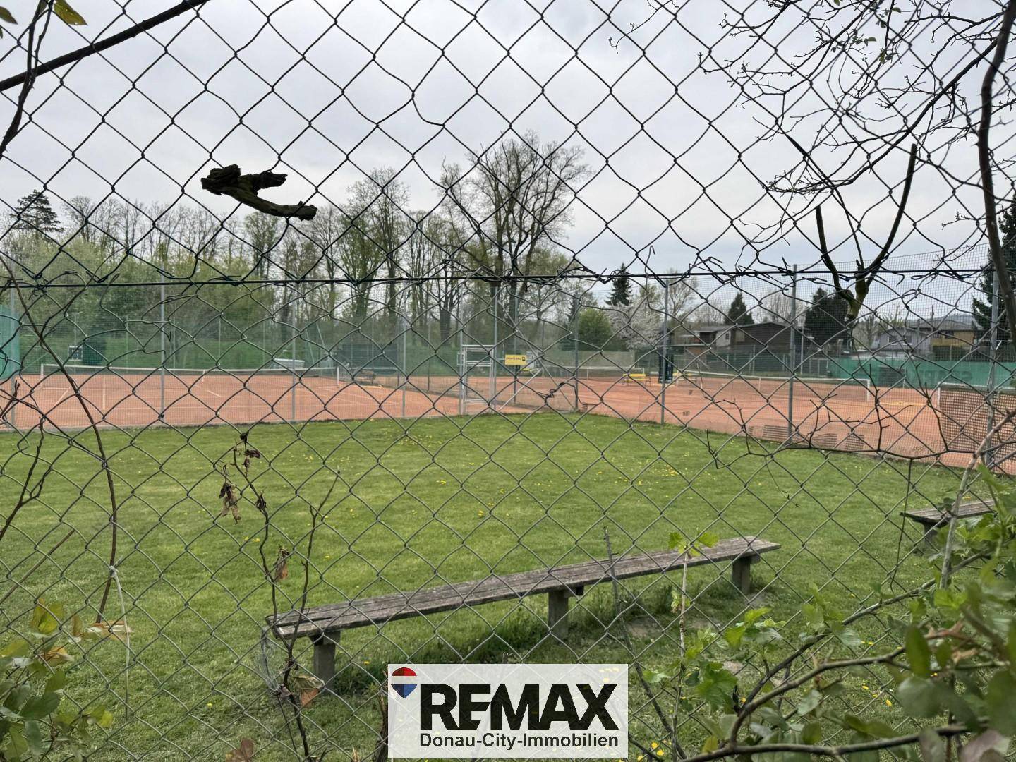 Blick durch einen Zaun auf zwei Tennisplätze mit rotem Sand und Sitzbänken.