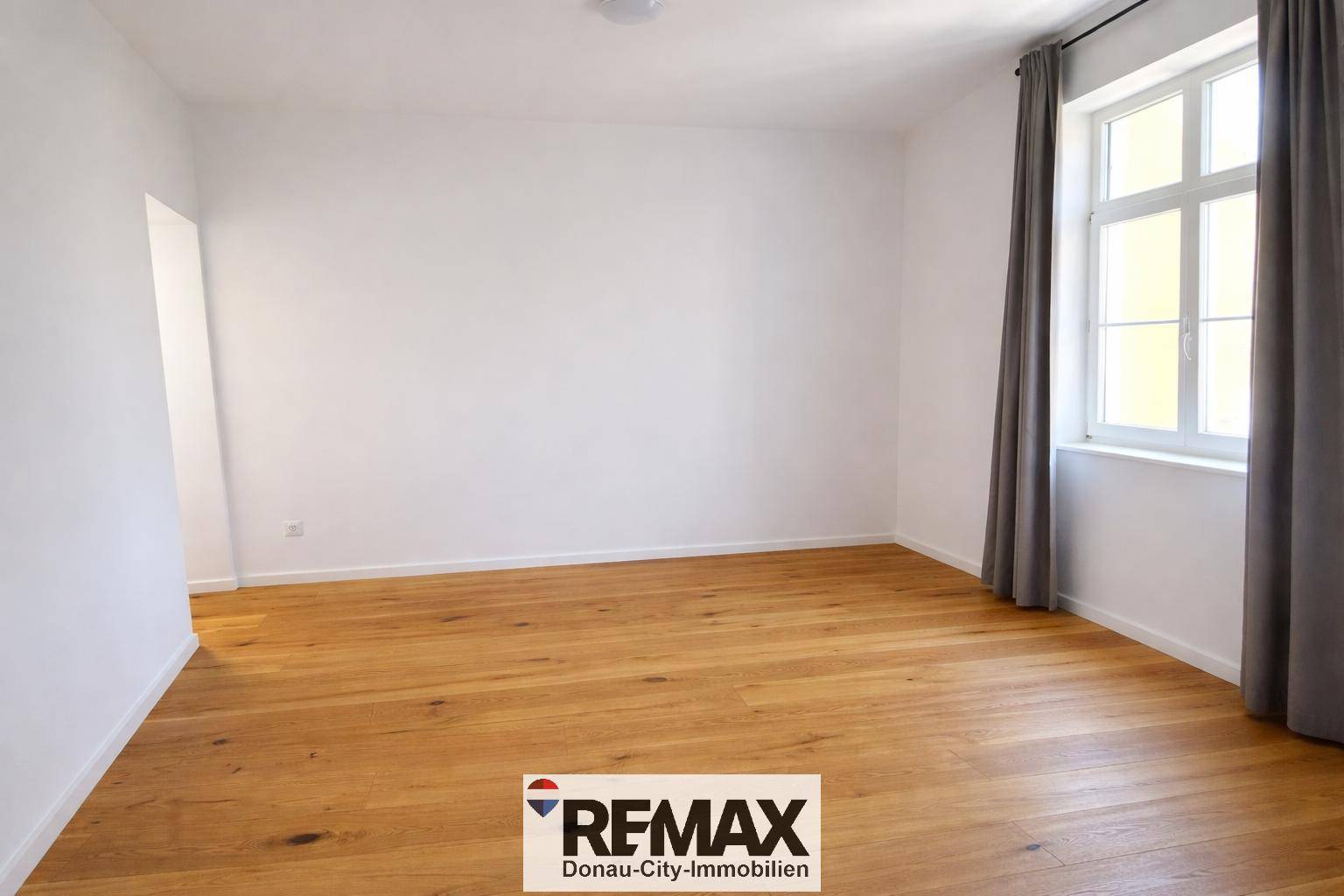 Heller, leerer Raum mit Holzboden, Fenster und Vorhängen.