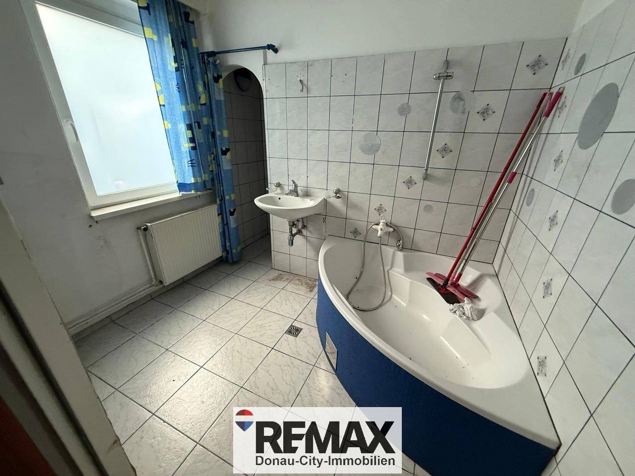 Badezimmer mit Fliesenboden, Eckbadewanne und Fenster, das Tageslicht hereinlässt.