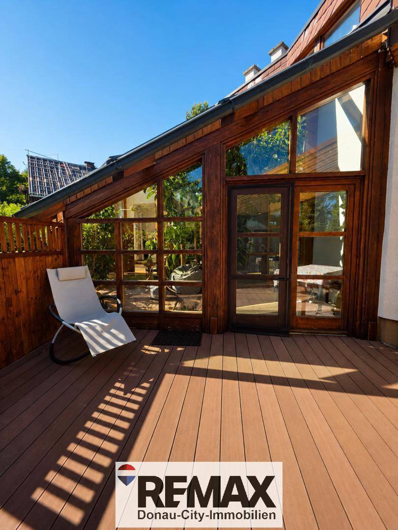 Sonnige Holzterrasse mit Liegestuhl und direktem Zugang zum Wintergarten, umgeben von viel Grün.