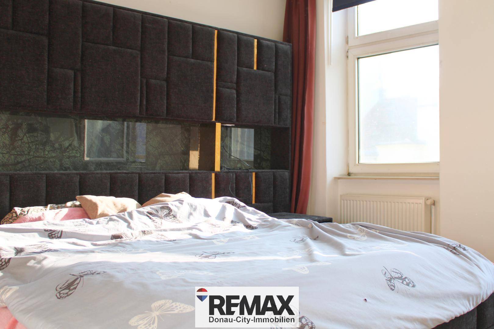 Schlafzimmer mit großem Bett, gepolstertem Kopfteil und einem Fenster mit rotem Vorhang.