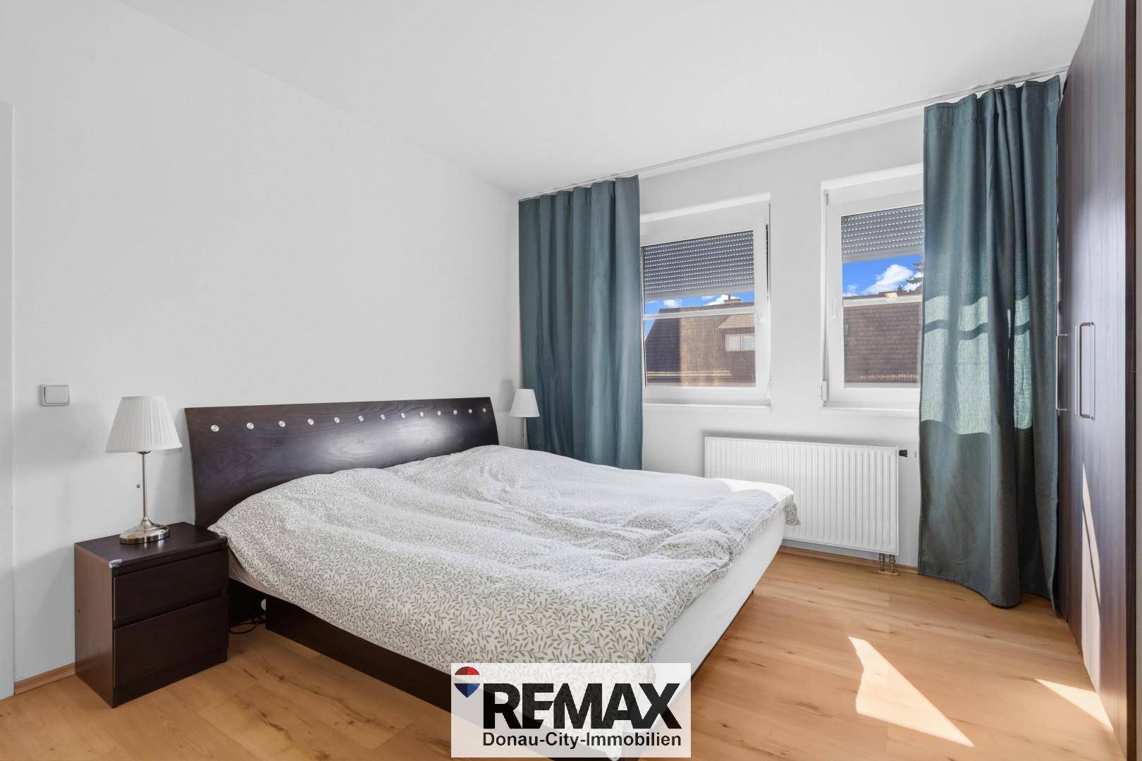 Schlafzimmer mit Doppelbett, Holzboden und zwei Fenstern mit Blick nach draußen.