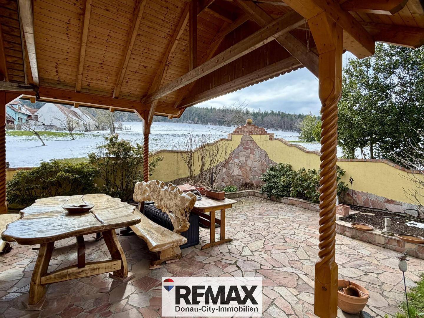 Überdachte Terrasse mit rustikalen Holzmöbeln und Steinboden, umgeben von einer winterlichen Landschaft.