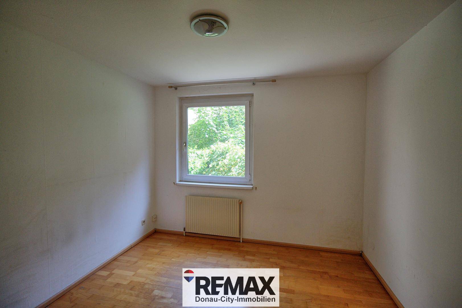 Helles Schlafzimmer mit Parkettboden und Fenster, das einen ruhigen Blick ins Grüne ermöglicht.