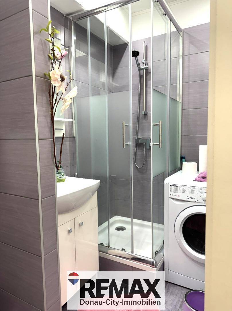 Helles Badezimmer mit Duschkabine, Waschbecken und Waschmaschine in grauen Fliesen.