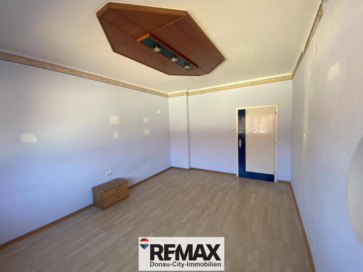 Geräumiges Zimmer mit hellem Laminatboden, dekorativer Decke und Tapetenbordüre an den Wänden.