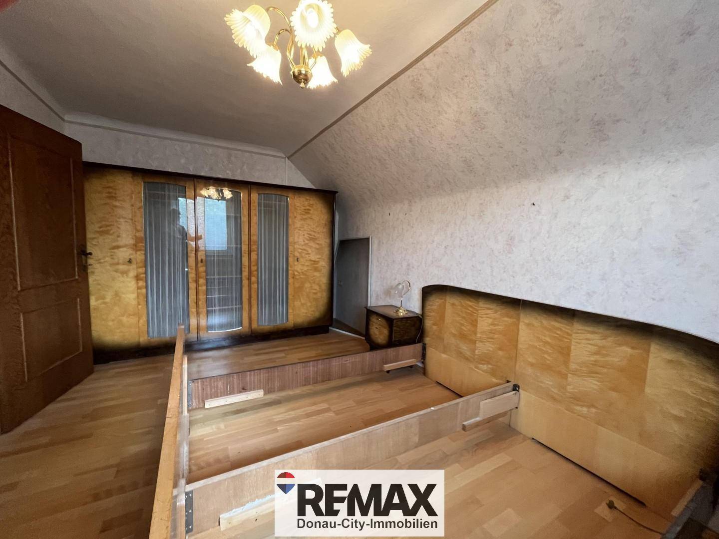 Schlafzimmer mit Bettrahmen, großem Holzschrank und gemusterter Tapete, mit Dachschräge.