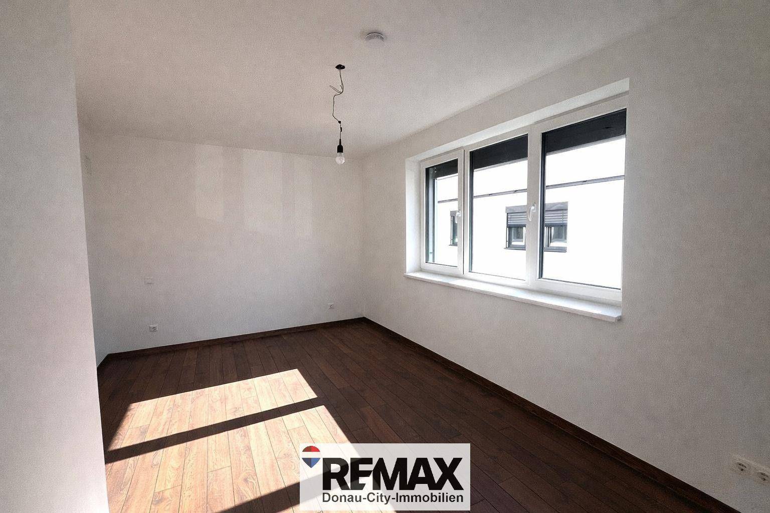 Helles Zimmer mit dunklem Holzboden und großem Fenster, das viel Tageslicht hereinlässt.