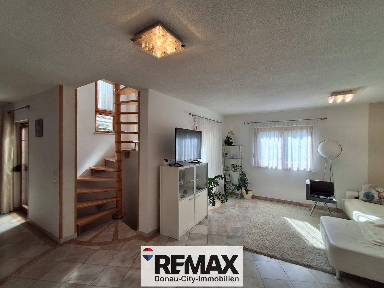 Helles Wohnzimmer mit Fliesenboden, weißem Sofa und einer eleganten Holzwendeltreppe zum Obergeschoss.