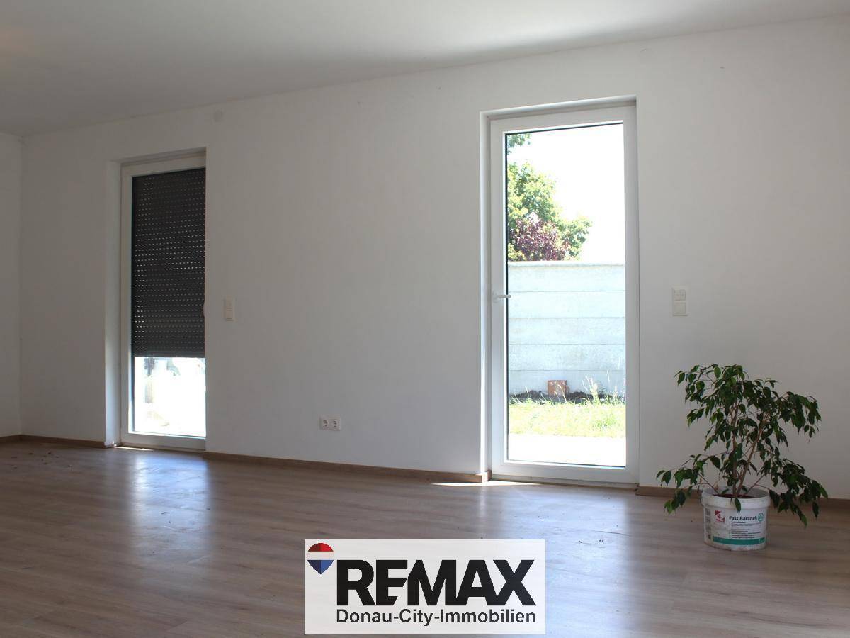 Lichtdurchfluteter Wohnraum mit großen Fenstern, einer davon mit Rollladen, und Laminatboden.