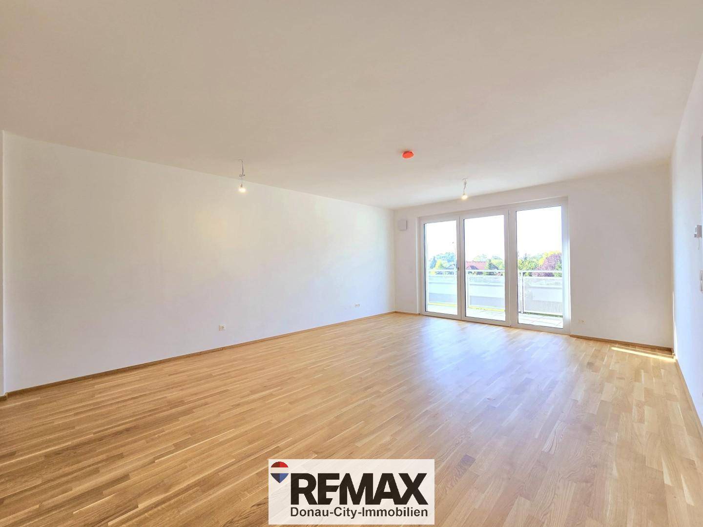 Geräumiges, leeres Wohnzimmer mit hellem Holzboden und großen Fenstern zum Balkon.