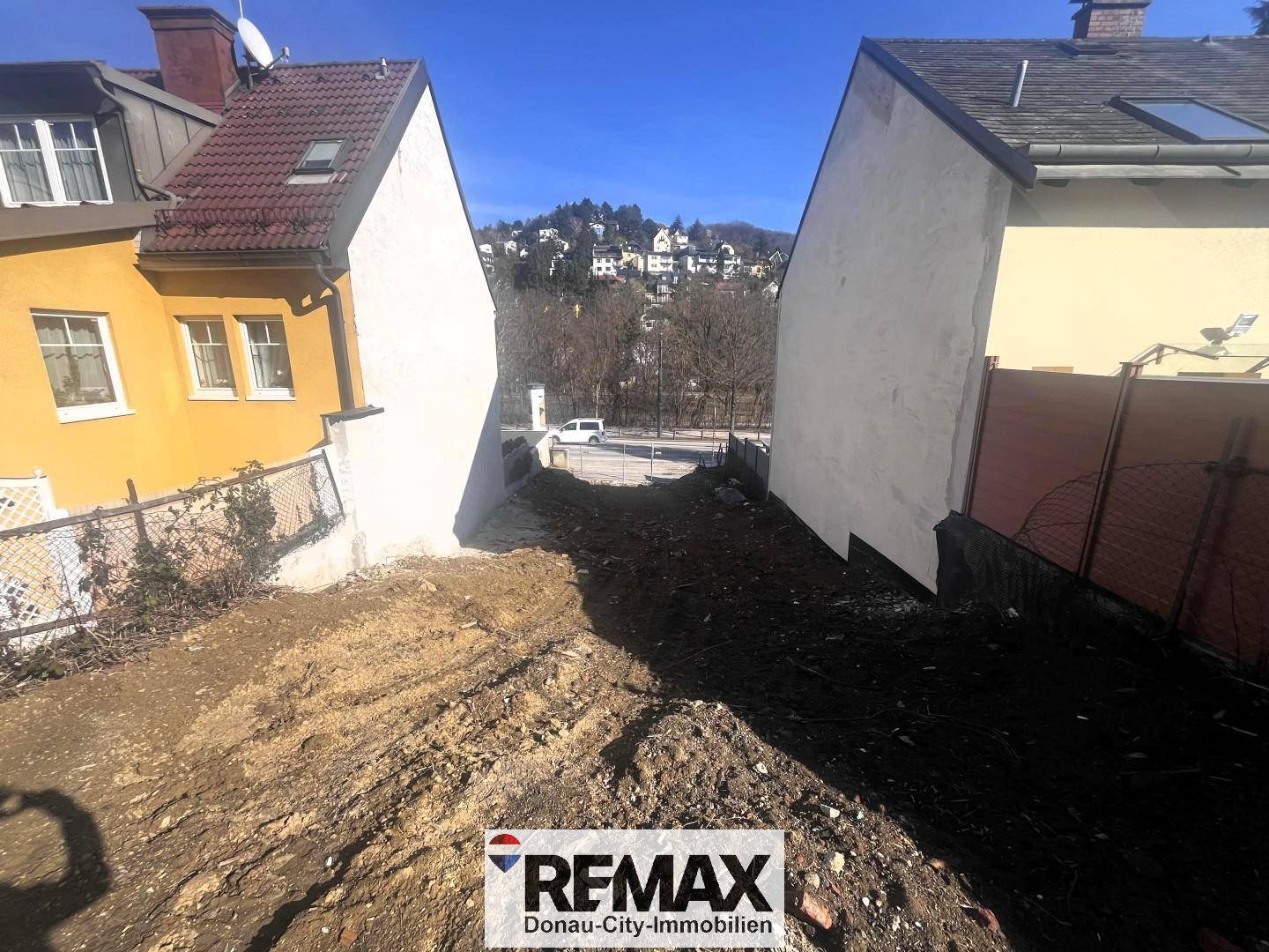Unbebautes Hanggrundstück zwischen zwei Häusern mit Blick auf eine Straße und bewaldete Hügel.
