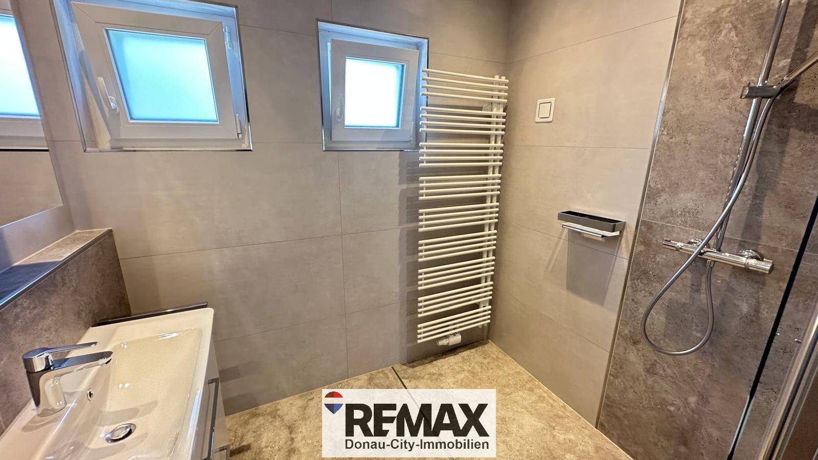 Modernes Badezimmer mit Dusche, Handtuchheizkörper und zwei kleinen Fenstern.