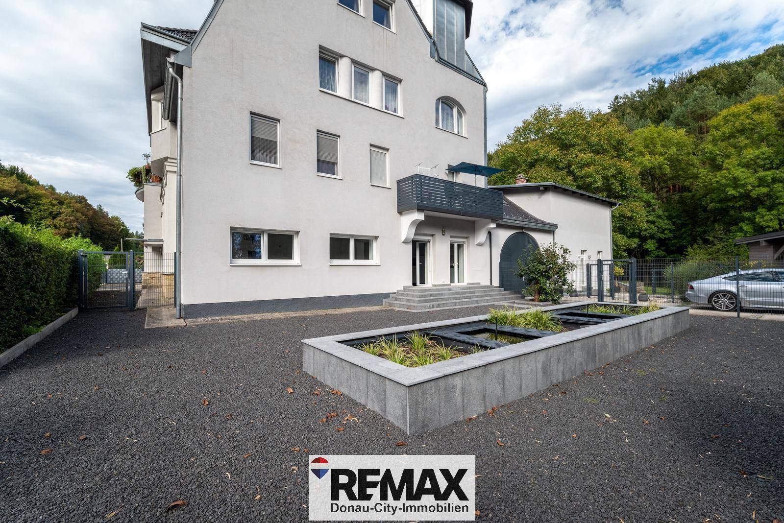 Seitenansicht des Gebäudes mit Kiesauffahrt und einem modernen Hochbeet mit Pflanzen.