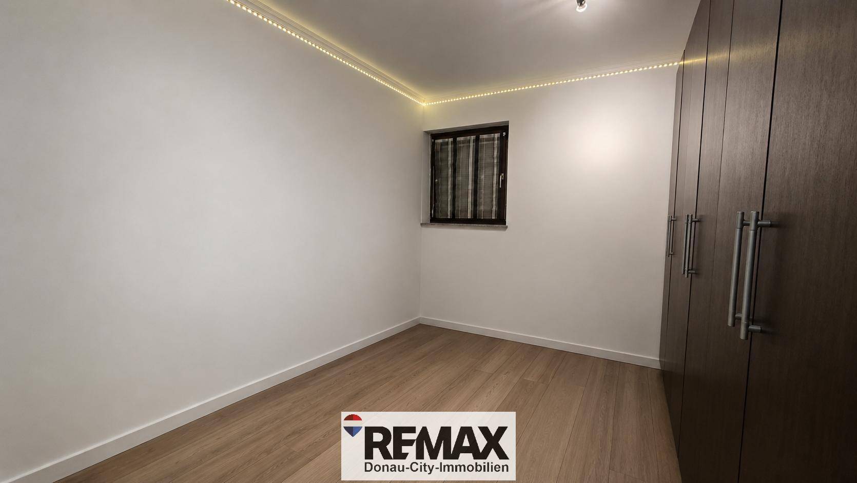 Helles Zimmer mit großem Einbauschrank, Fenster und indirekter Deckenbeleuchtung.