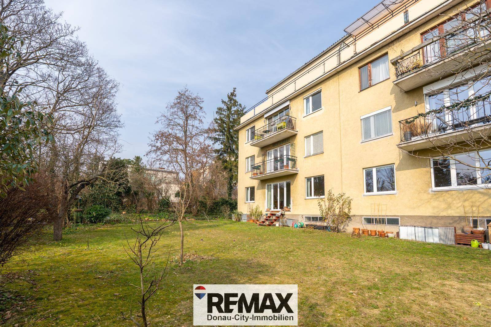 Gepflegter Gartenbereich mit Rasen und Bäumen vor einem Mehrfamilienhaus mit Balkonen.
