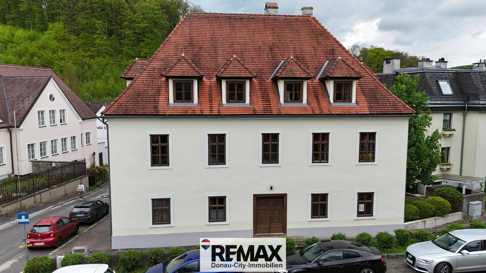 Historisches Gebäude mit traditionellem Ziegeldach und mehreren Gaubenfenstern in grüner Umgebung.