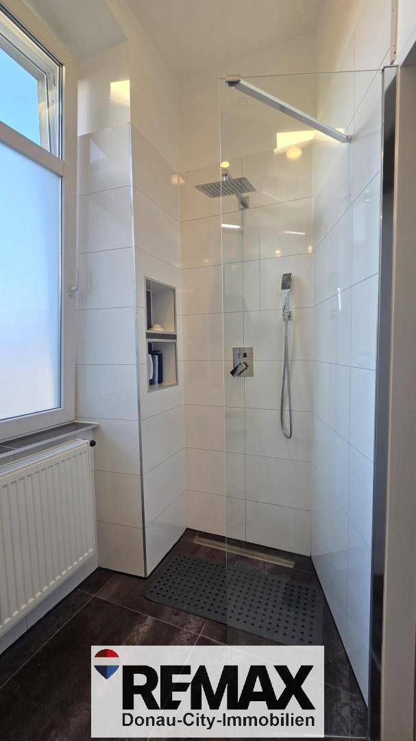 Helle Dusche mit Glasabtrennung, Regenduschkopf und Fenster für natürliches Tageslicht.