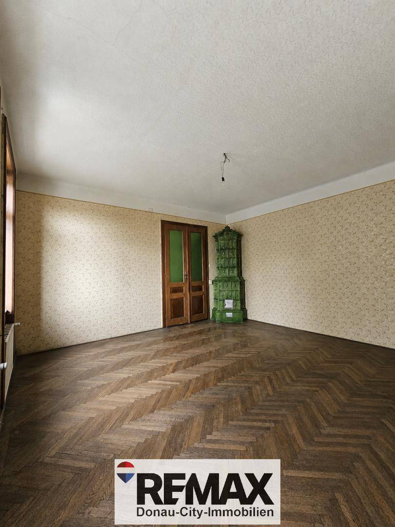 Geräumiges Zimmer mit traditionellem Kachelofen, Fischgrätparkett und Holztüren, bereit zur Renovierung.