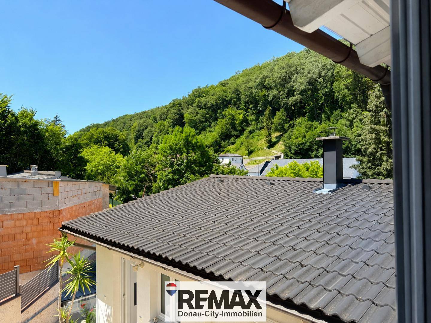 Blick über ein Dach mit Schornstein auf die umliegende grüne Hügellandschaft.