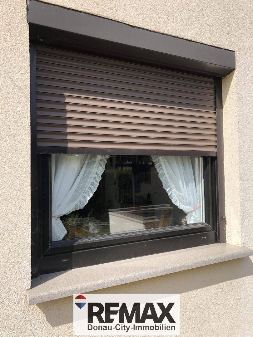 Fenster mit geschlossenem braunen Rollladen und weißer Gardine.