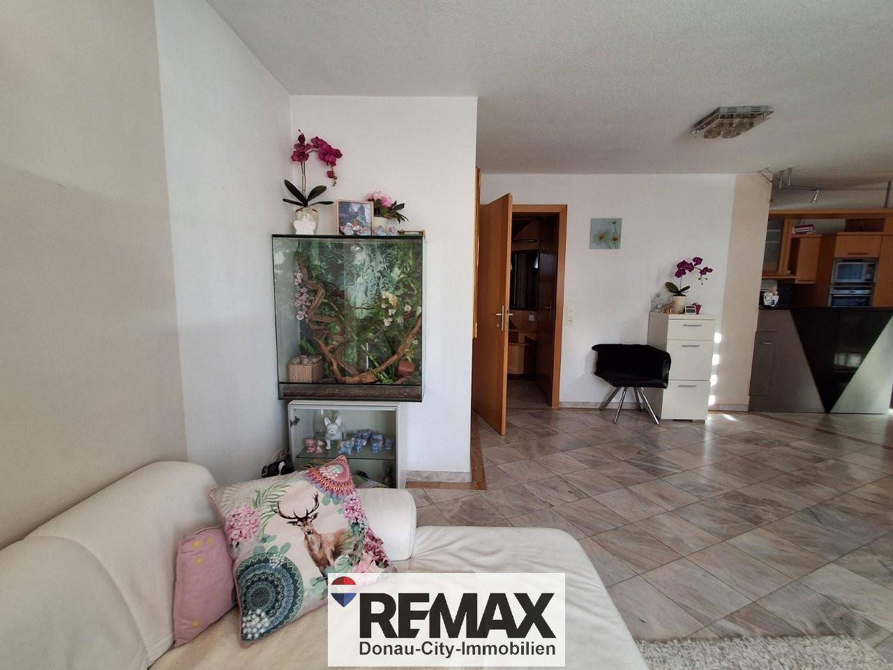 Wohnbereich mit hellem Sofa, einem Aquarium und offenem Übergang zur Küche, mit Fliesenboden.